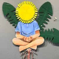 A wall-mounted cardboard sculpture painted with acrylics. It depicts a child in a light blue button-up shirt and khaki shorts sitting cross-legged with its hands cupped together. In the place of a head, there is a dandelion flower, and three dandelion leaves extend behind the figure. There is a long brown root trailing down underneath the figure, and floating to the right is a single dandelion seed